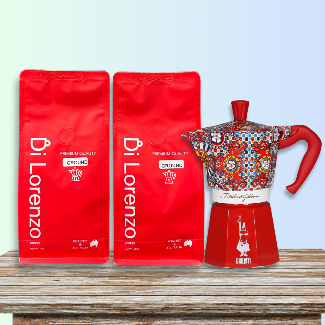 Bright red bags of Di Lorenzo premium quality ground coffee, ground specifically for Moka stove top, beside a colorful, artistically designed Bialetti moka pot on a rustic wooden table, against a soft blue gradient backdrop.