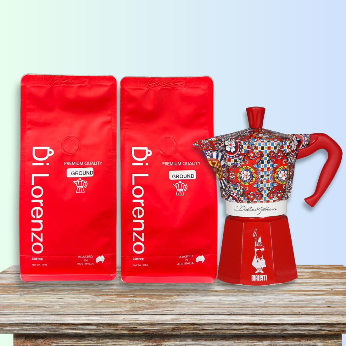Bright red bags of Di Lorenzo premium quality ground coffee, ground specifically for Moka stove top,  beside a colorful, artistically designed Bialetti moka pot on a rustic wooden table, against a soft blue gradient backdrop.