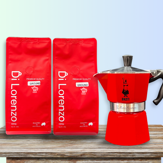 Twin red bags of Di Lorenzo premium quality ground coffee flank a classic grey Bialetti red Moka  stovetop espresso maker, all presented on a textured wooden surface with a gradient blue sky background.