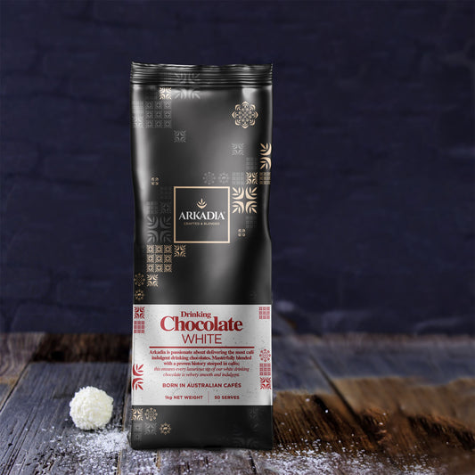 One packet of white drinking chocolate with a black and gold bag, in a natural ingredient set with wooden board and sprinkle of coconut flakes pared with a cup of dil lorenzo coffee